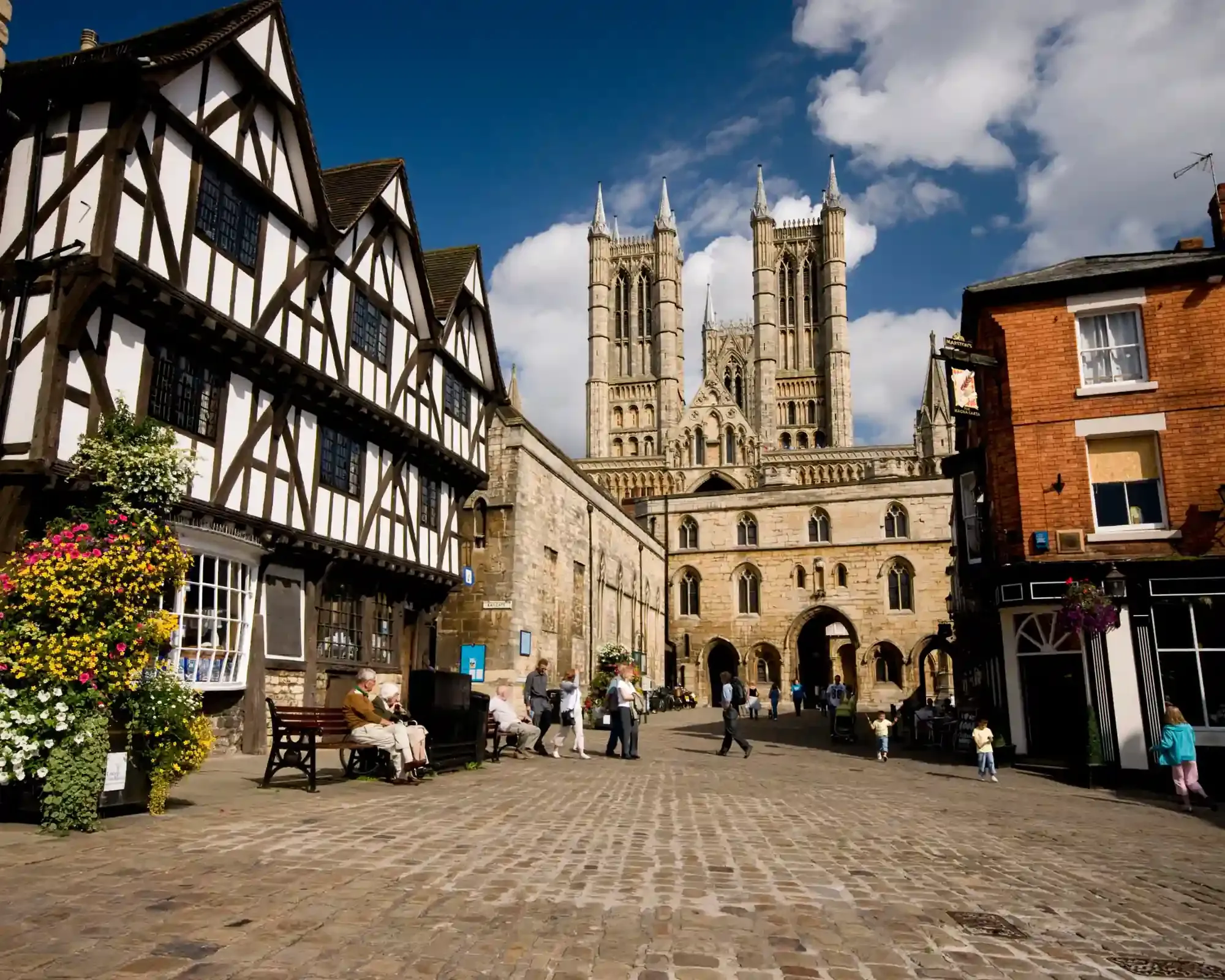 Castle Square in Lincoln, with Tudor timber-framed buildings, people walking on cobbles and Lincoln Cathedral's towers behind the Exchequer Gate.