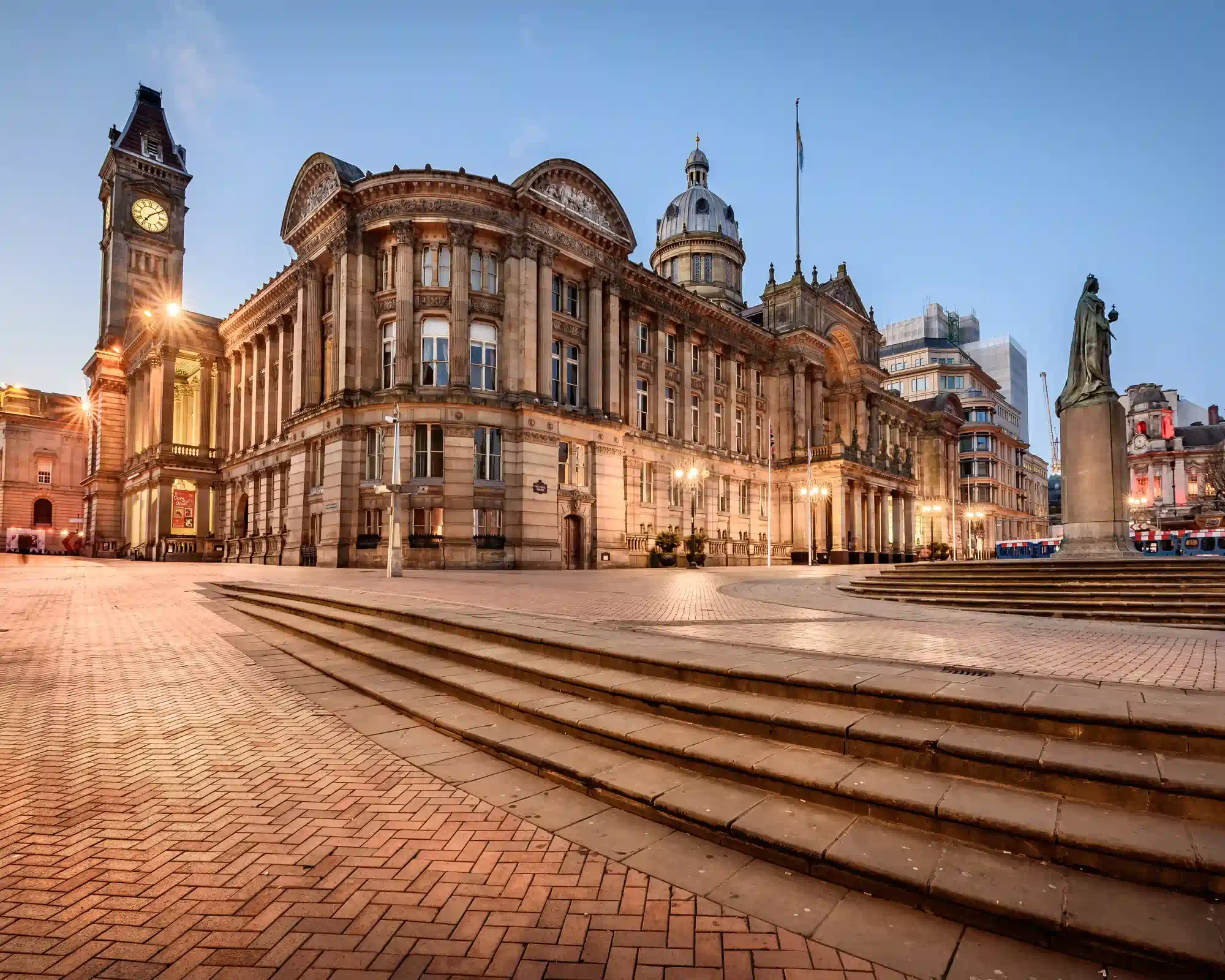 Birmingham Council House and its clock tower lit up at dusk in Victoria Square, with a statue of Queen Victoria and wide stone steps in front.