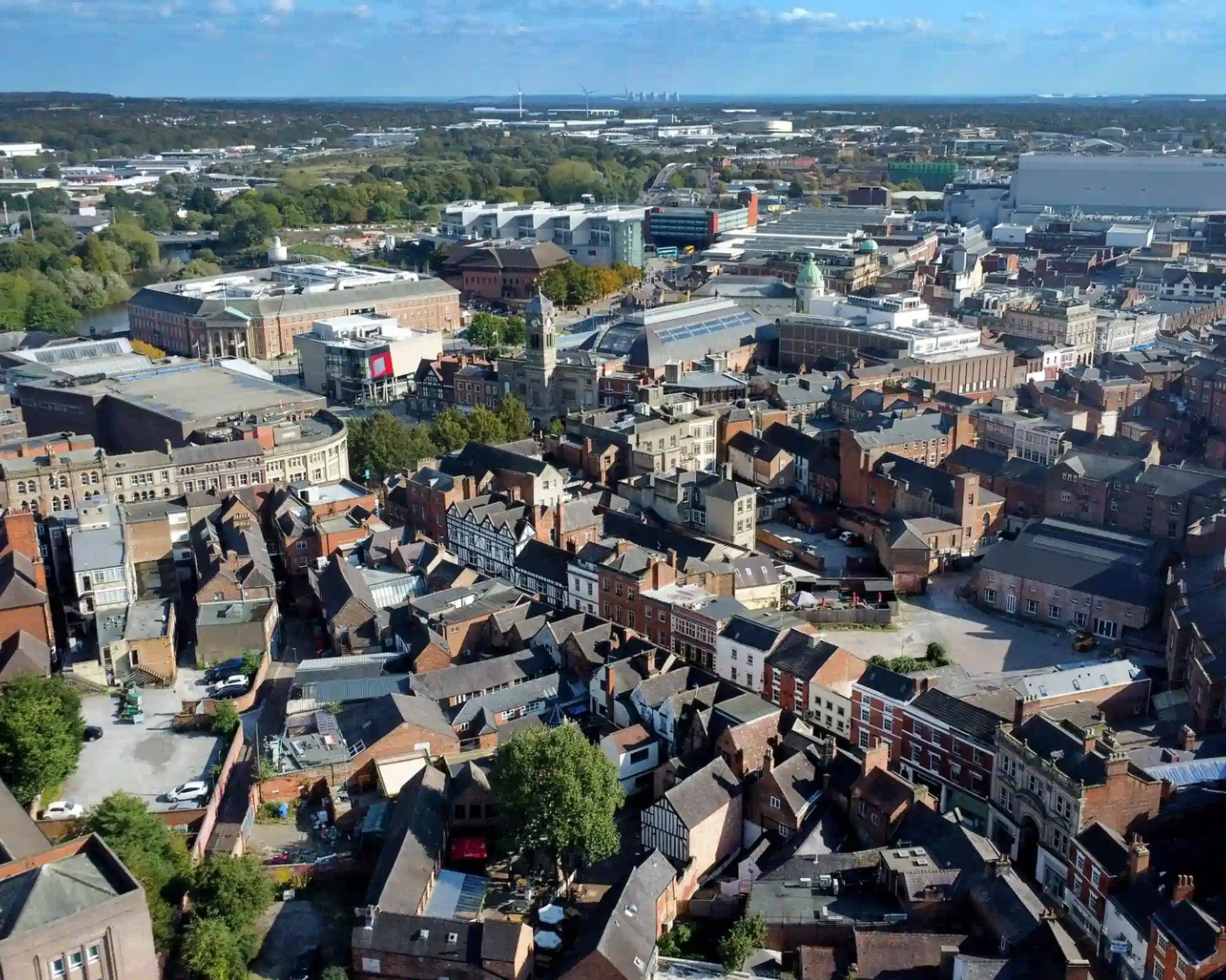 Aerial view of Derby city centre on a sunny day, with the Guildhall clock tower rising above red-brick rooftops, the Council House beside the River Derwent, and the city stretching to the horizon.