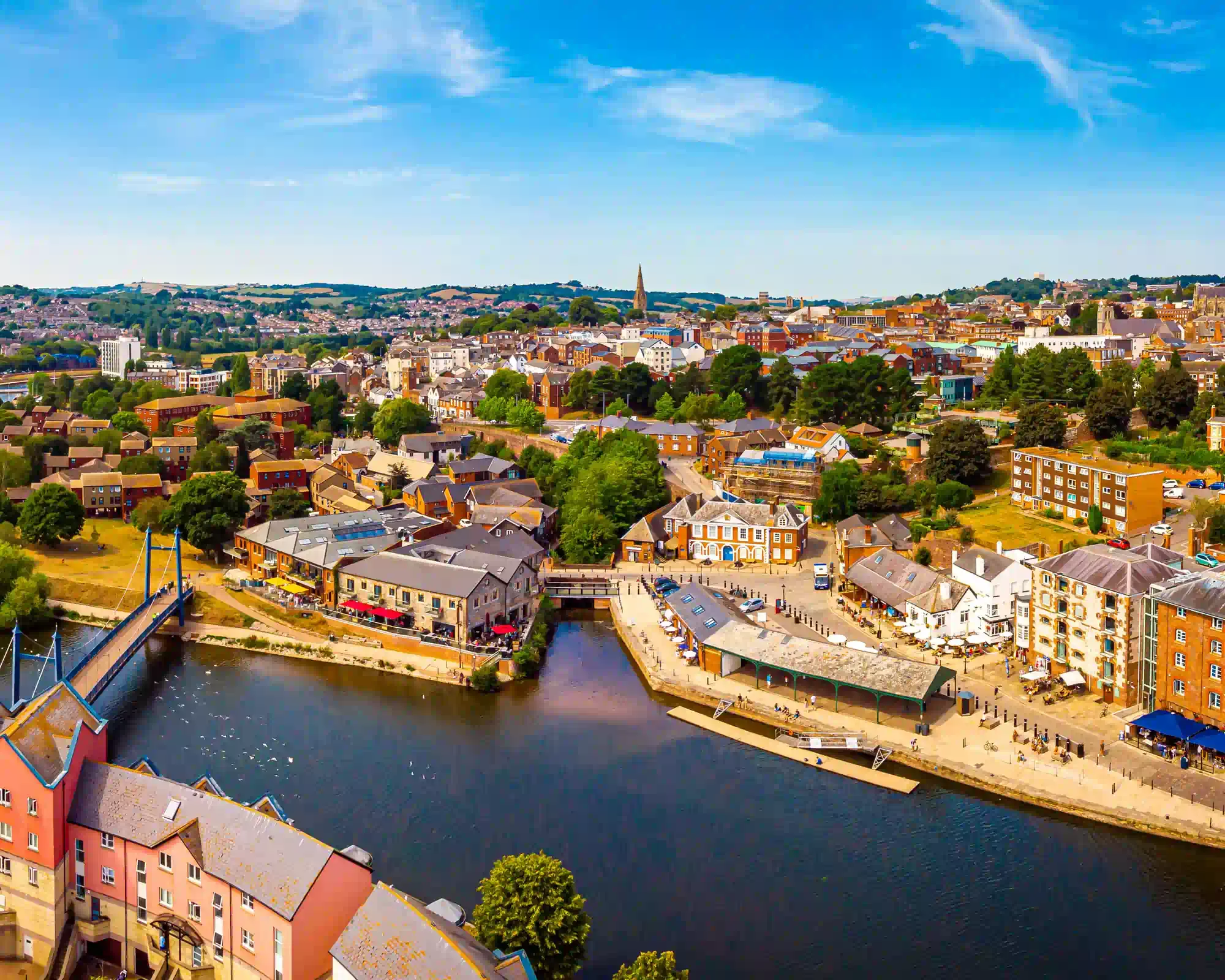 Aerial view of Exeter Quayside on a sunny day, with the River Exe curving past historic warehouses, riverside cafés and red-brick buildings, and the city rising behind.