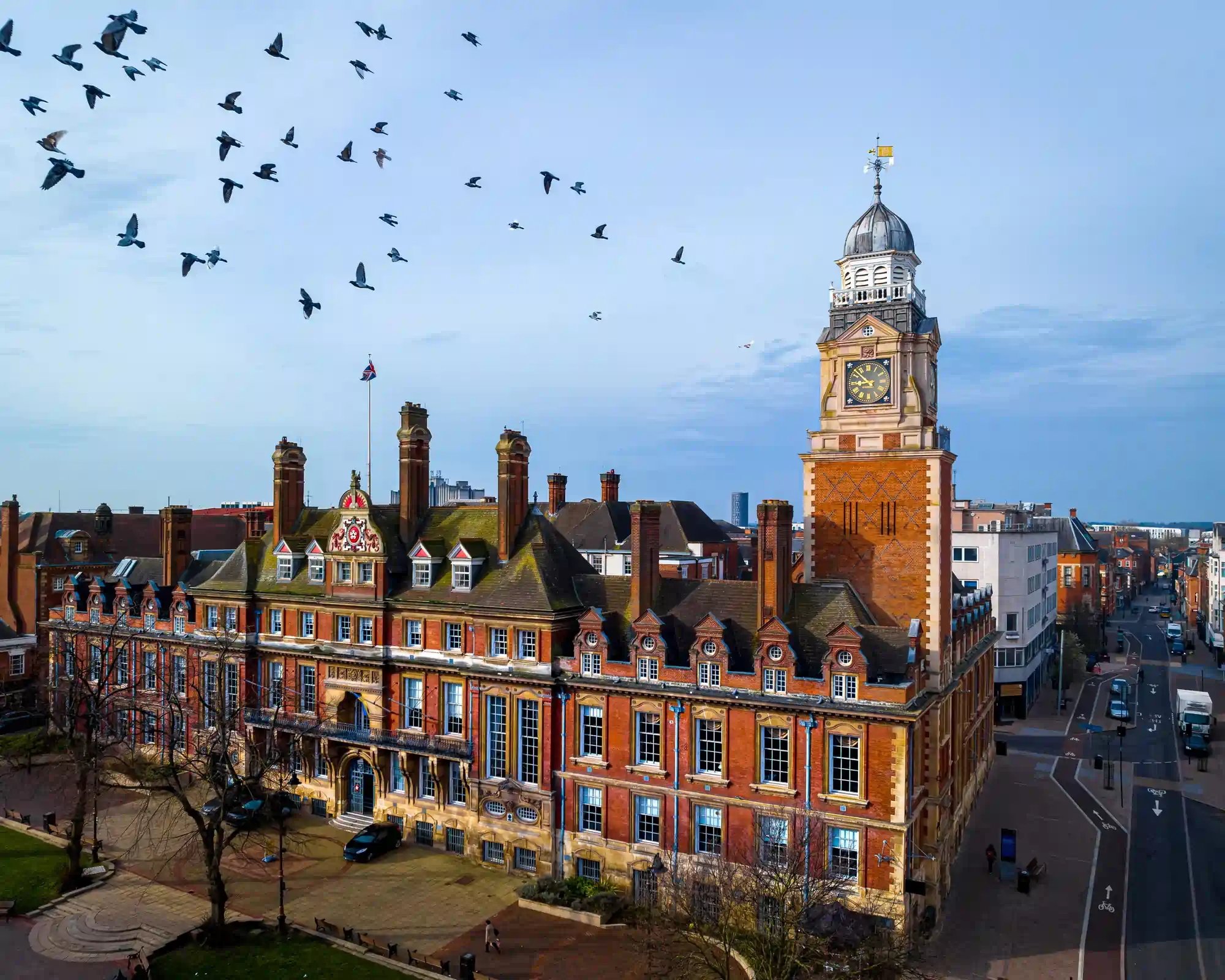 Leicester Town Hall, a red-brick Victorian building with a clock tower, with pigeons flying across a blue sky above Town Hall Square.