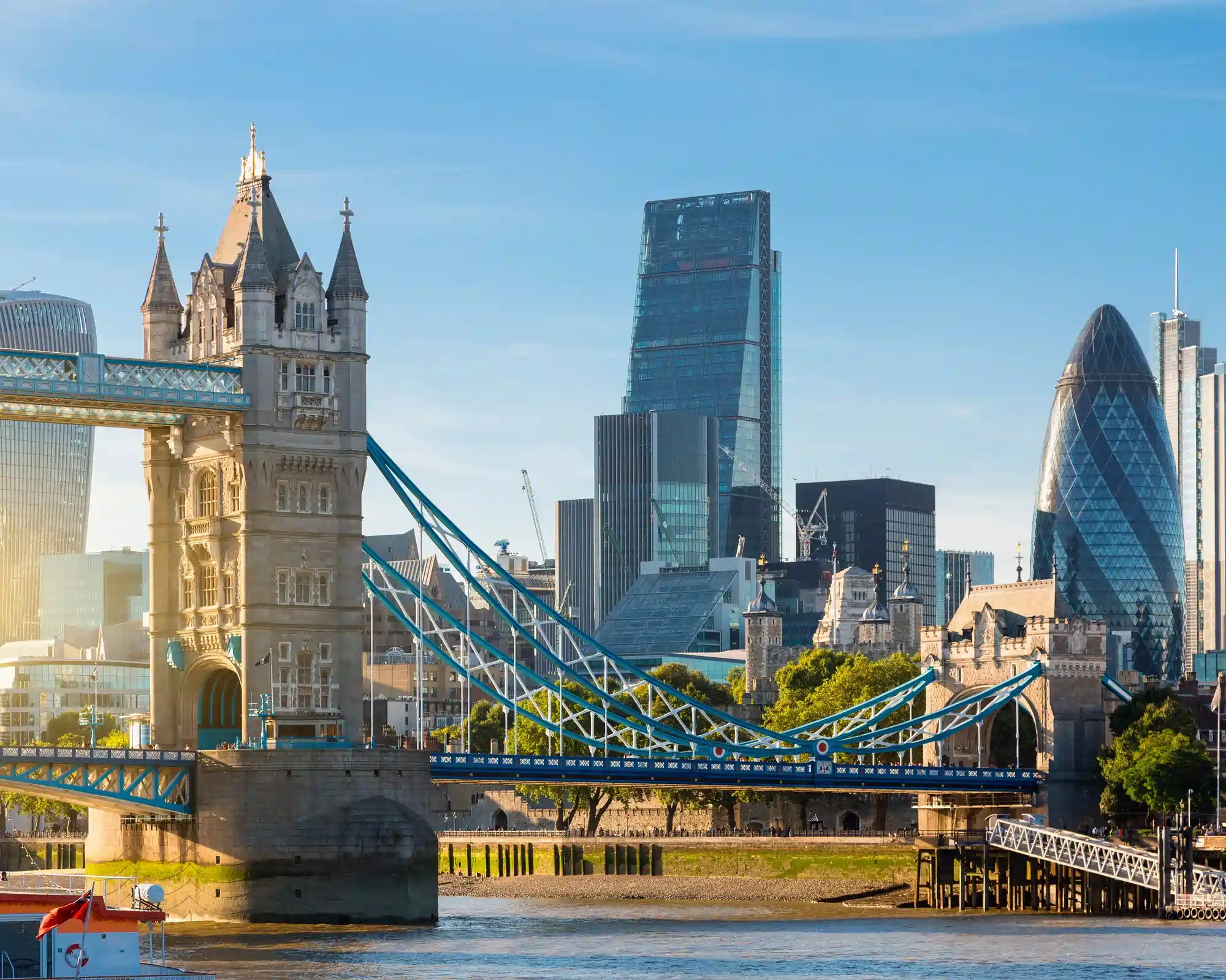 Tower Bridge over the River Thames on a sunny day, with the Tower of London and City skyscrapers including the Gherkin behind.