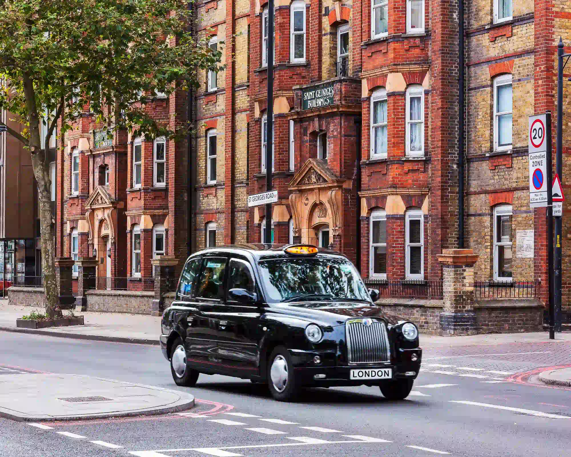 A black London taxi driving past Saint George's Buildings, a Victorian red-brick apartment block on St George's Road.