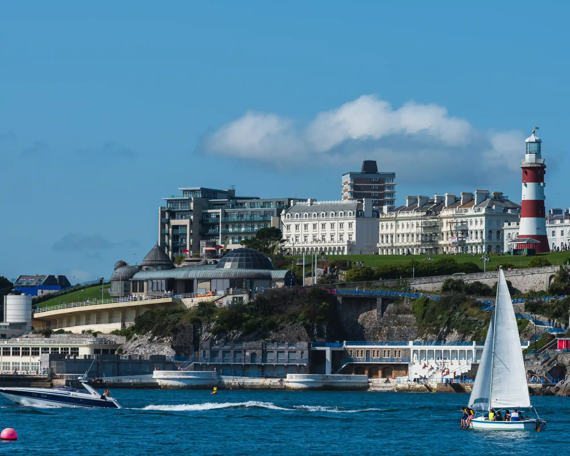 Plymouth Hoe on a sunny day, with Smeaton's Tower lighthouse and white seafront buildings above the water, and a sailing boat and speedboat on Plymouth Sound.