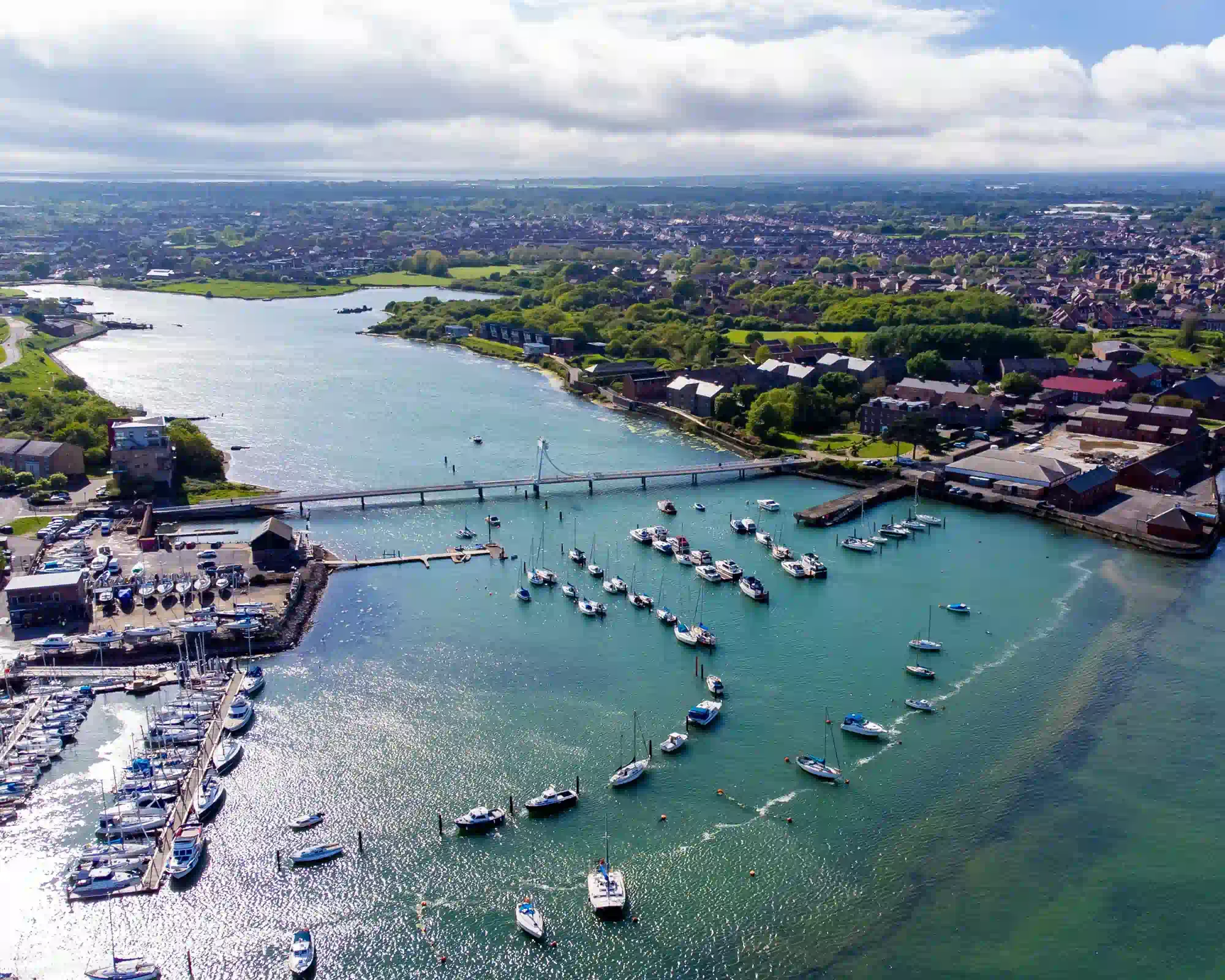 Aerial view of a harbour marina on a sunny day, with sailing boats moored on turquoise water, a cable-stayed footbridge across a creek, and red-brick buildings and houses beyond.