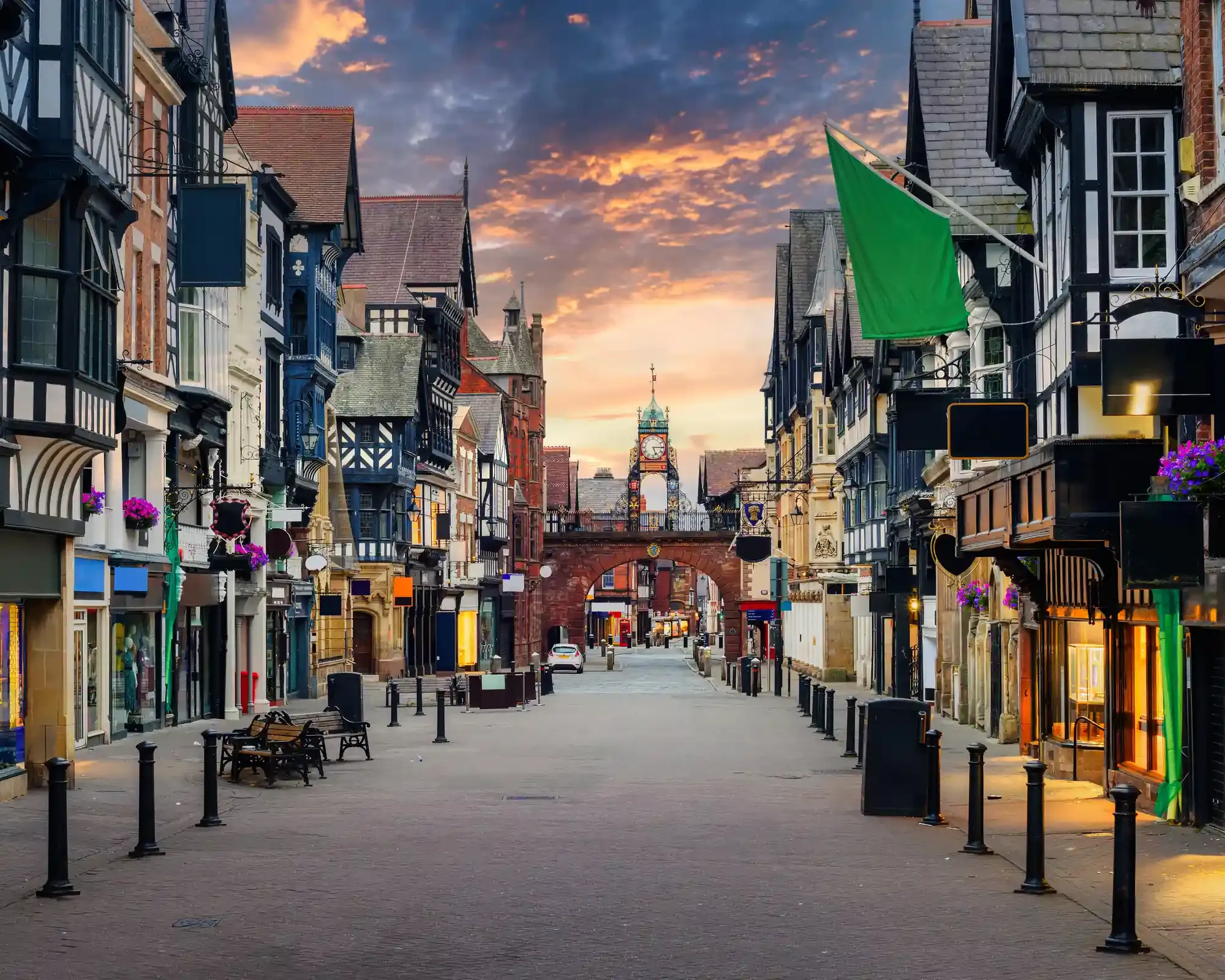 Black-and-white timber-framed shops along Eastgate Street in Chester at sunset, with the Eastgate Clock on the city walls ahead.