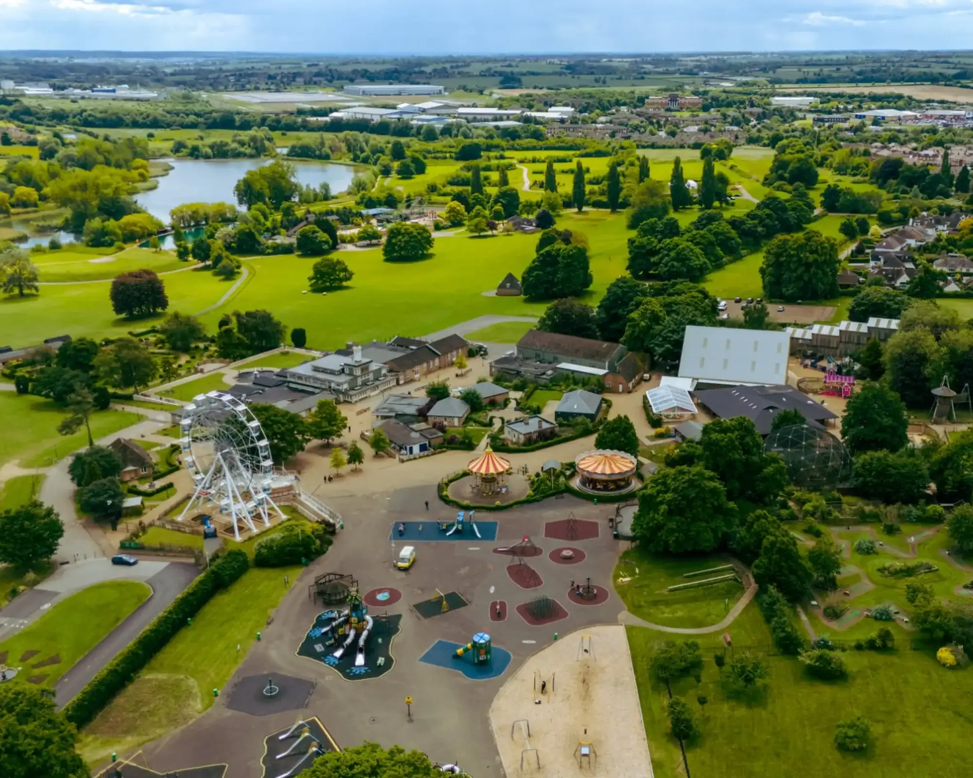 Aerial view of Wicksteed Park in Kettering, with its big wheel, carousels, playground and lake, and housing and countryside beyond.