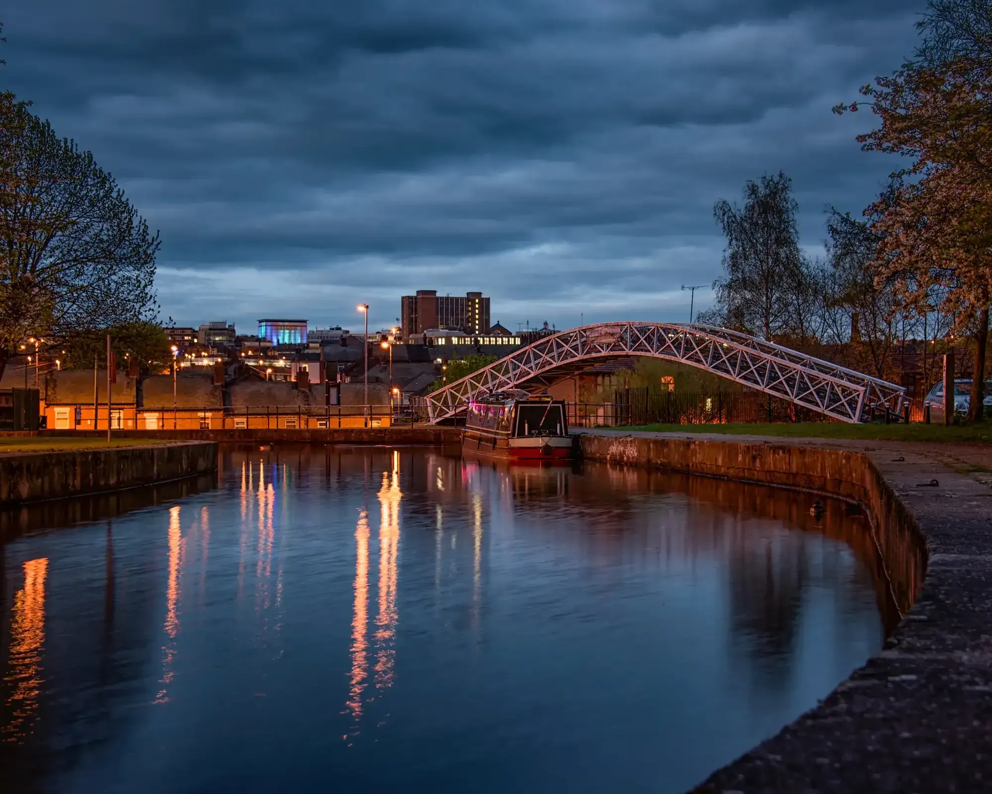 A canal footbridge in Stoke-on-Trent at dusk, with the city skyline reflected in the water
