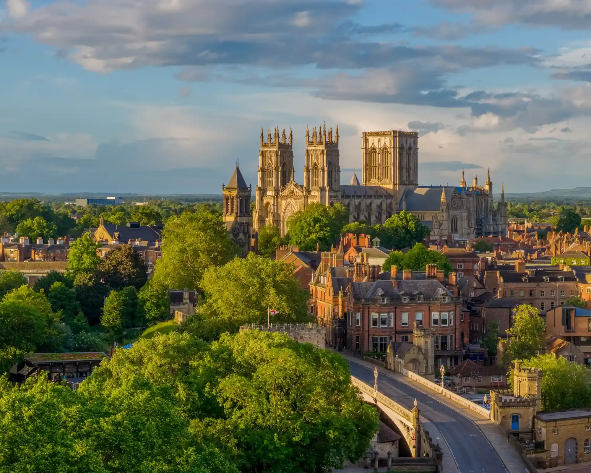 York Minster rising above the rooftops in evening sunlight, with Lendal Bridge and the city walls in the foreground.