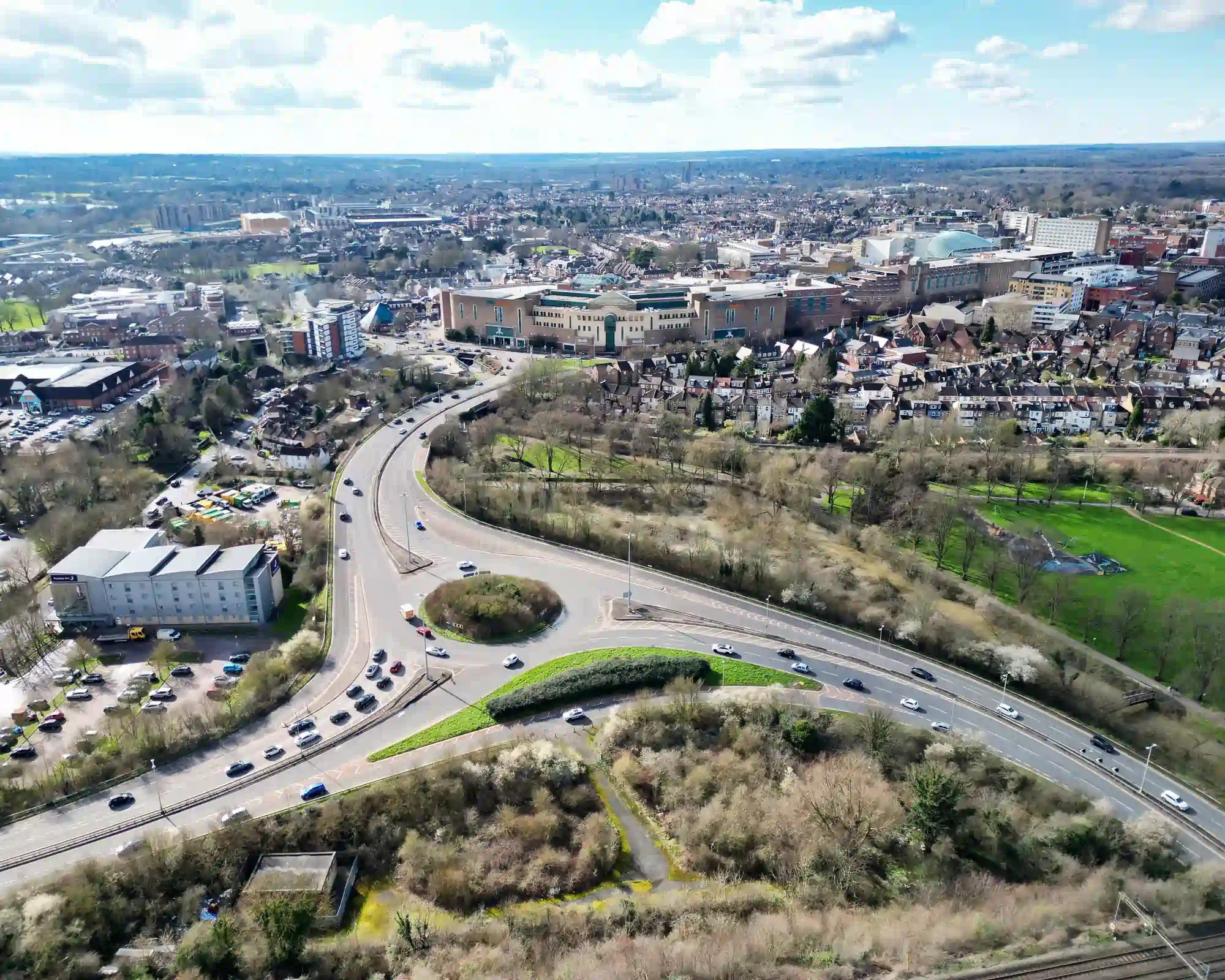 Aerial view of Watford on a sunny day, with a busy roundabout and a curving road leading to the town centre shopping area, and green parkland and rows of houses beside it.