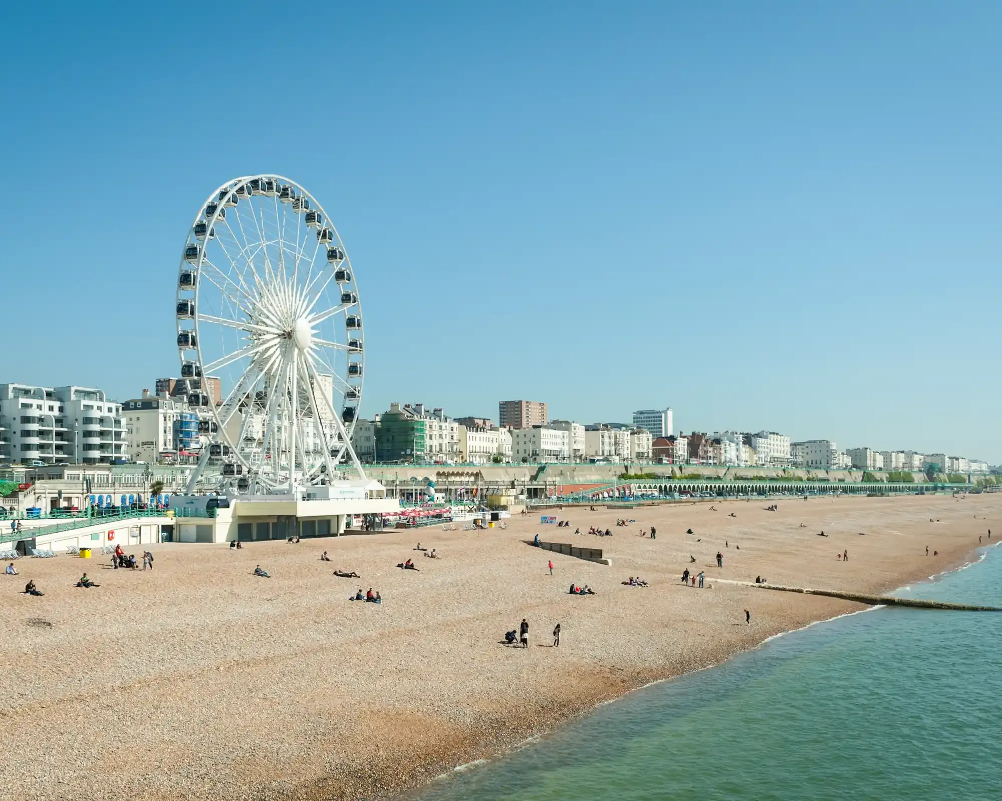 Brighton beach on a sunny day, with the Brighton Wheel and white seafront buildings beside a pebble beach and calm sea.